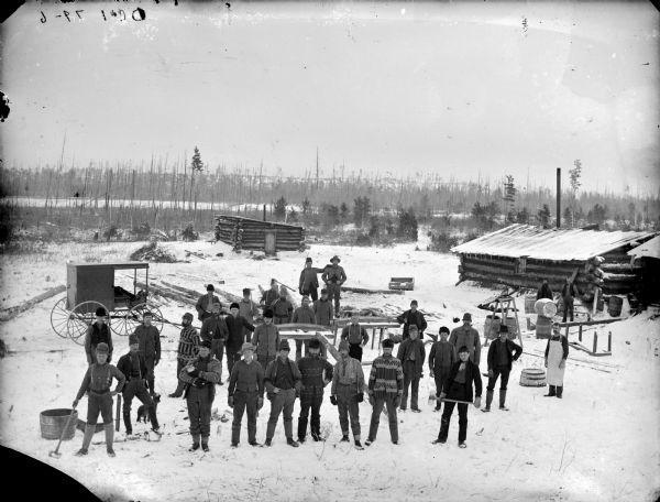 Elevated view of men posed standing in a snow-covered logging camp. One man is holding a cat, and there is a dog standing nearby. The photographers wagon is in the background.