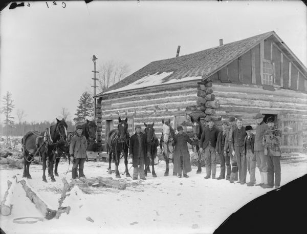 Group portrait of men posed standing on the snow-covered ground in front of a log building. Three men stand with teams of two horses.	