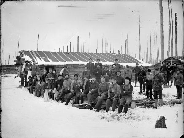 Group portrait of men, women. children and dogs posed standing, sitting and holding logging tools in a snow-covered logging camp in front of log buildings.