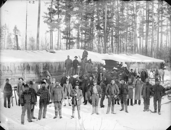 Elevated view of a group of men posed standing and holding logging tools in snow-covered logging camp in front of log buildings.	