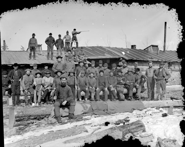 Group of men posed holding logging tools while sitting and standing in front of and atop a log building.