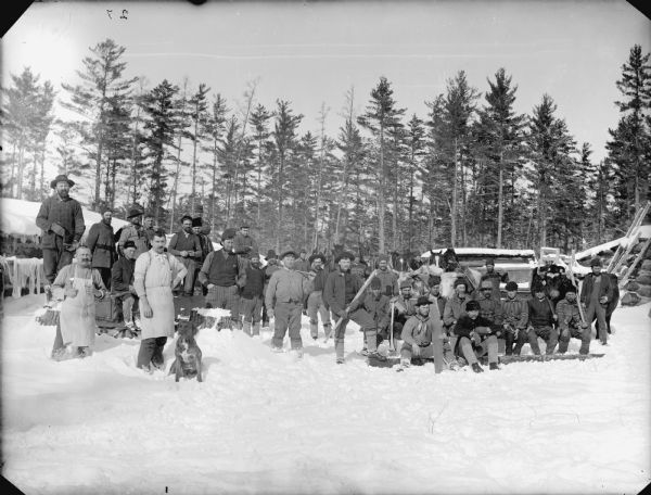 Group of men posed holding logging tools while sitting and standing on the snow-covered ground in front of a log building and the woods.	