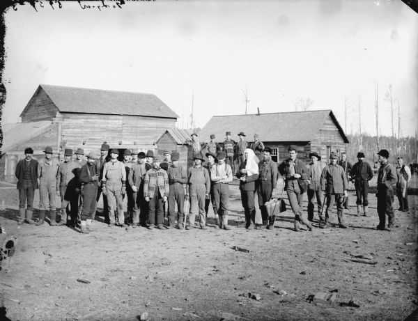 Group of men posed standing in front of two wooden buildings, possibly a logging camp.	