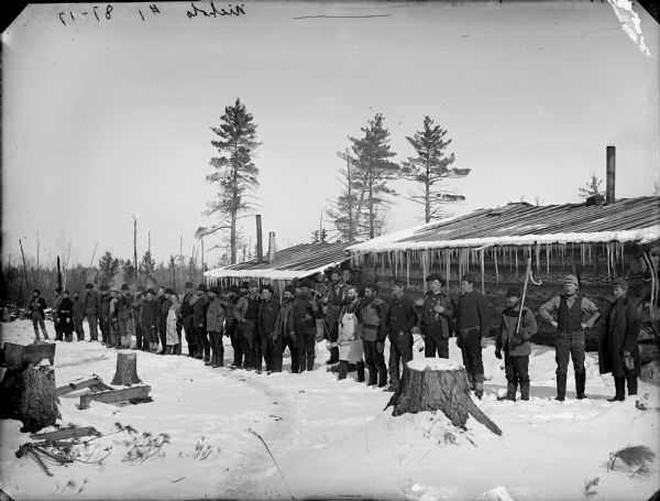 Group of men posed standing and holding logging tools while standing in a line on the snow-covered ground in front of two log buildings.