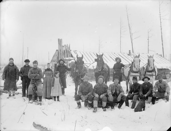 Group of men, women, children and a dog posed sitting and standing in front of a log building in the snow. Two men are holding two teams of two horses.