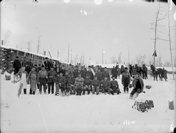 Group of men posed standing and sitting on benches on the snow-covered ground in front of log buildings and several horse teams.	