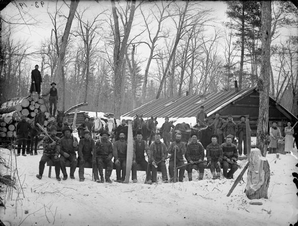 Group portrait of men posed holding logging tools, while standing and sitting on log benches. There is snow on the ground. On the left behind the group is a sled loaded with logs. On the right is a log building.