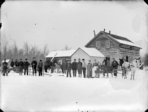 Group of men posed holding logging tools. Three men stand with six horses. The group is standing on the snow-covered ground in front of a log building. Two women stand by a horse-drawn sleigh with two girls sitting inside.