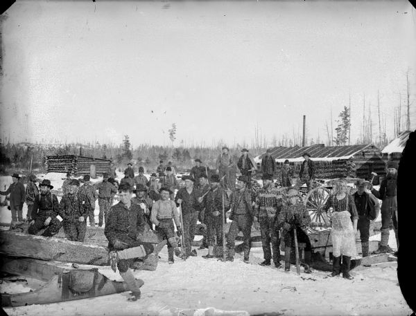 Group of men posed sitting and standing and holding logging tools in front of log buildings.	There is snow on the ground.
