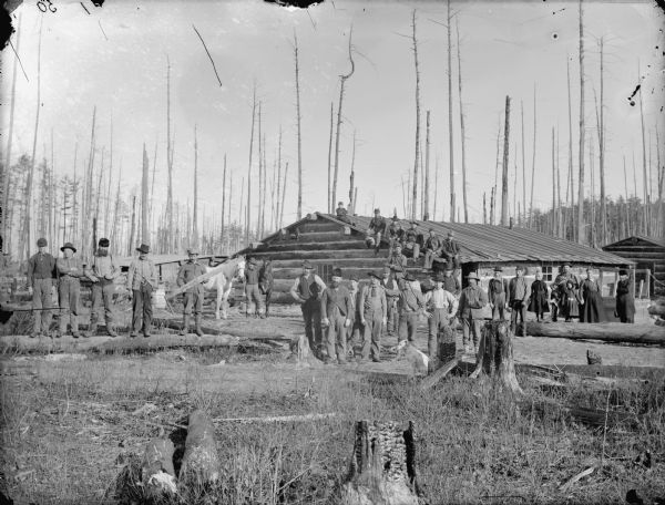 Group of men posing and holding logging tools while standing and sitting in front of, and on top of, a log building. In the background on the right is a woman and four children.	