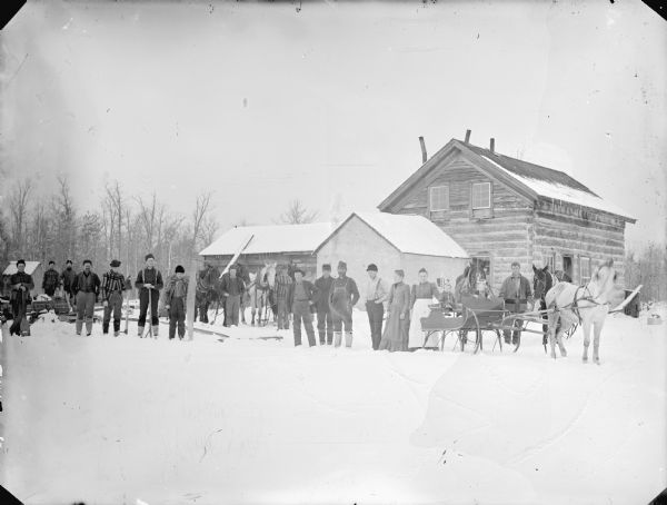 Group of men posed holding logging tools. Three men stand with six horses. The group is standing on the snow-covered ground in front of a log building. Two women stand by a horse-drawn sleigh with two girls sitting inside.	