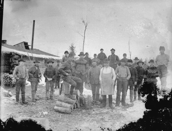 Group of men posing standing on the snow-covered ground in front of a log building. In the foreground one man is sitting on a pile of logs.