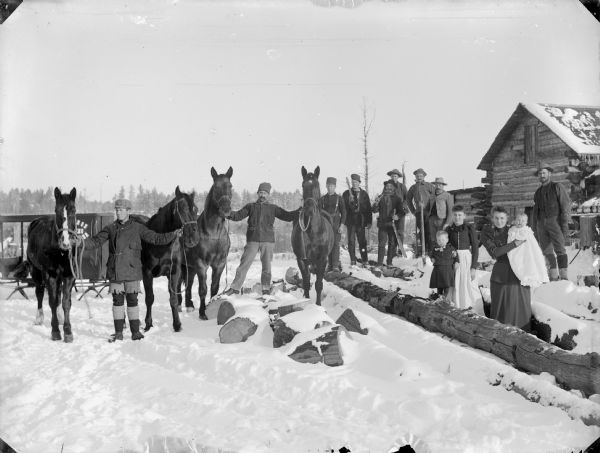 Group of men, women and children standing on the snow-covered ground in front of a log building. Some of the men are posed holding logging tools, and two of the men stand holding the reins of teams of horses. Two women stand with two young children. The photographers wagon of C.R. Monroe is in the background on the left.	