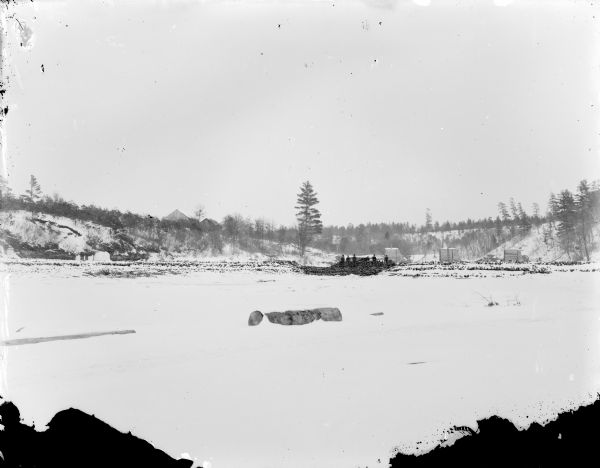 View over snow-covered ground or a frozen river of a group of men sitting on piles of logs. Behind them on the right is a log sorting pier. There are two buildings on a hill in the background on the left.