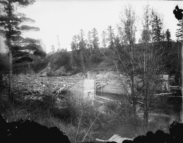View from shoreline of two men posed standing on log sorting piers half a mile above the Railroad Bridge on the Black River. There is a large log jam behind them, and the far shoreline has steep cliffs.