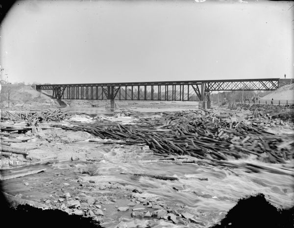 View from shoreline of a log jam under a railroad bridge about half a mile from the Black River Falls Dam. On the opposite shoreline, men sit atop a fence. Another man stands at the top of the hill near the railroad tracks.