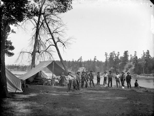Group of men and a boy posed holding logging tools and standing in front of tents on the banks of a river, probably just south of Black River Falls. A man wearing an apron and holding a coffee pot stands near an open-sided tent with a long table and benches set with dishes for meals. The group is possibly a clean-up crew for stray logs. A man on the far right sitting on the ground is wearing a top hat and smoking a pipe. In the far background on the right is a bridge and buildings.	