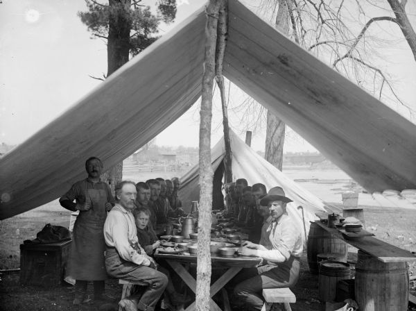 Group of men and one boy posed sitting on benches on either side of a long table for a meal in an open-sided tent on the banks of a river. In the background are buildings and a bridge over the river. A man wearing an apron, perhaps the cook, stands at the left of the group.