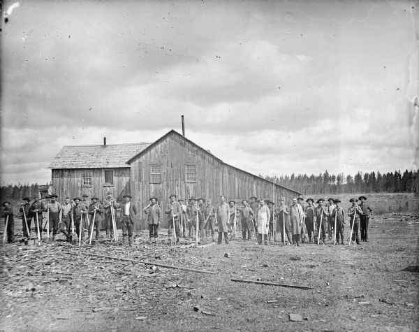Group portrait of men holding logging tools posed standing in front of a wooden building.	