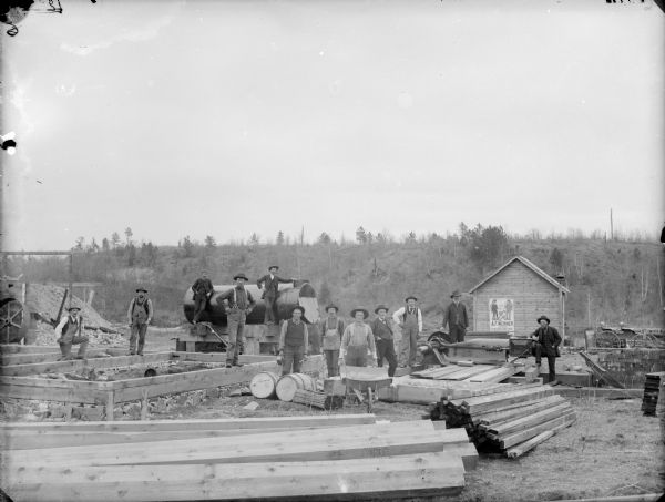 Men posed standing at the construction site. They are probably laying the foundation of a building. A small wooden building in the background has a poster that says: "Fine Stock Boys' Clothing A.F. Werner."