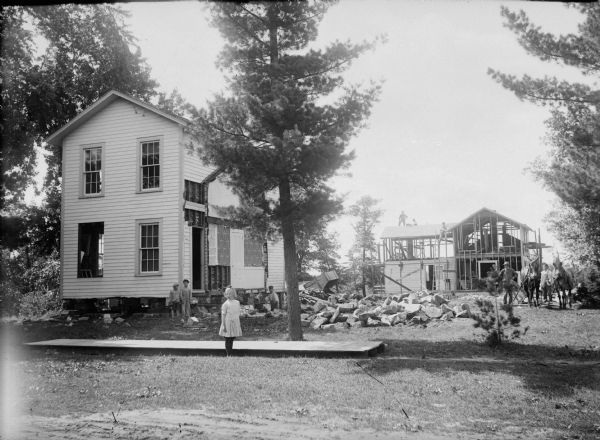 Men posed demolishing one house and building another on the same property, possibly the R.P. Ravey property. Two boys and a girl are posed standing in the foreground.