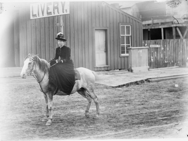 Mrs. Charles Van Schaick, posing on horseback, is riding past the livery stable. It was unusual for women at this time to ride horseback.