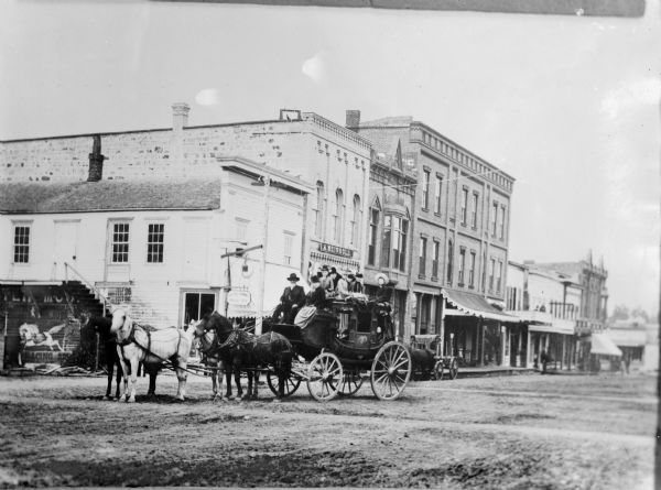 Men and women pose on an old stage coach pulled by four horses. In the background are commercial buildings at the corner of Main and First Streets. The coach originally belonged to Wisconsin state legislator W.T. Price, and in this photograph the driver is Ed Pratt. The heavily retouched print was made from an earlier negative by Charles Van Schaick.
