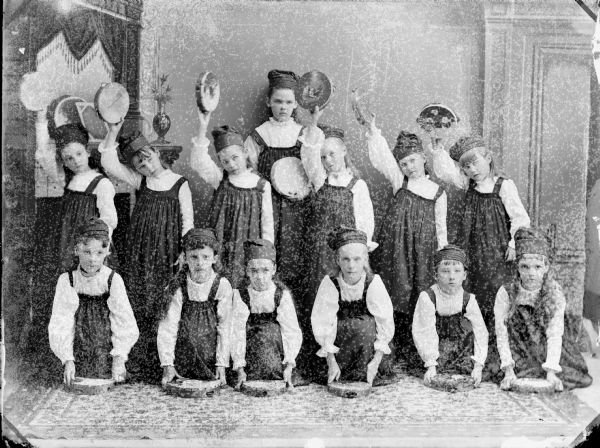 Studio portrait of thirteen girls in costume, posing sitting, standing, and holding tambourines in front of a painted backdrop.