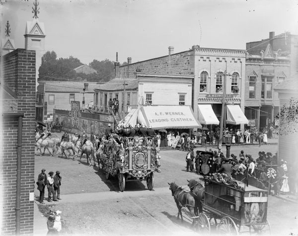 Elevated view of a crowd gathered at the intersection of First and Main Streets to watch a bandwagon and animal cage pass in a circus parade. Probably the Ringling Brothers Circus.	