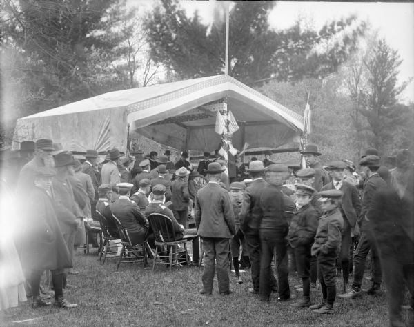 Crowd of men and boys gathered around a tent, probably listening to a political or patriotic speech or debate.	