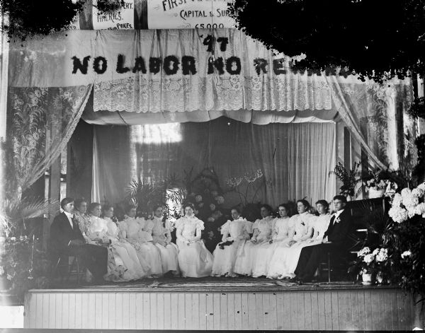 Two young men and ten young women in white dresses sitting in a semi circle on the stage of the Black River Falls Opera House. The stage is decorated with lace work curtains and floral arrangements, with the motto "No Labor, No Rewards" written above. Ruth Jones is sitting second from right.