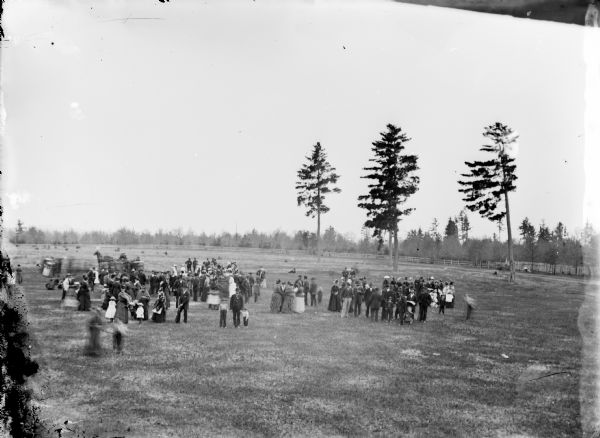 Elevated view of a large group of people gathered in an open field surrounded by a wooden fence.