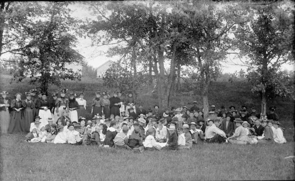 Large group sitting and standing among trees in a field. There are buildings in the far background.	