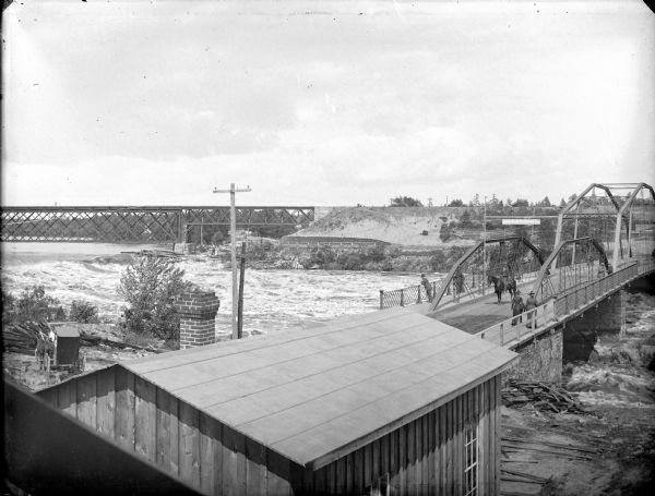 Elevated view over buildings of men and women walking across a bridge over a high river. There is a railroad bridge in the background.	