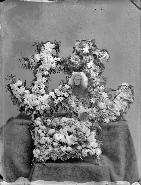 Studio portrait of memorial wreaths in the shape of anchors. The memorial includes a photographic portrait of a man, most likely John Best who died in 1890.