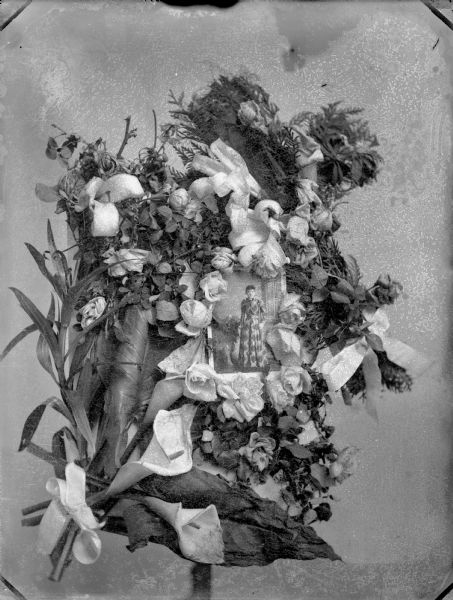 Studio portrait of a memorial flower wreath. In the middle is a full-length portrait of a woman in a dress.