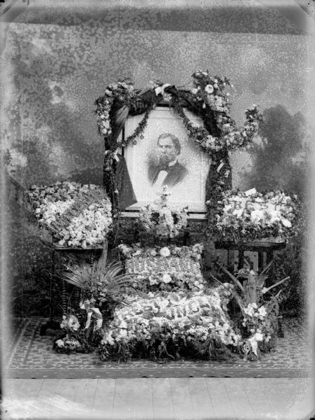 Studio portrait in front of a painted backdrop of memorial wreath grouping surrounding a draped portrait of W.T. Price. The wreaths have text which read: "Husband," "Rest," and "Rest Uncle".