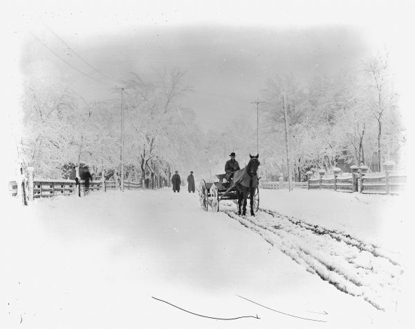 Winter scene with a man sitting in a wagon pulled by a single horse down a snow-covered road. The horse is wearing blinders and a turnout blanket. Two men stand in the road behind the wagon.	