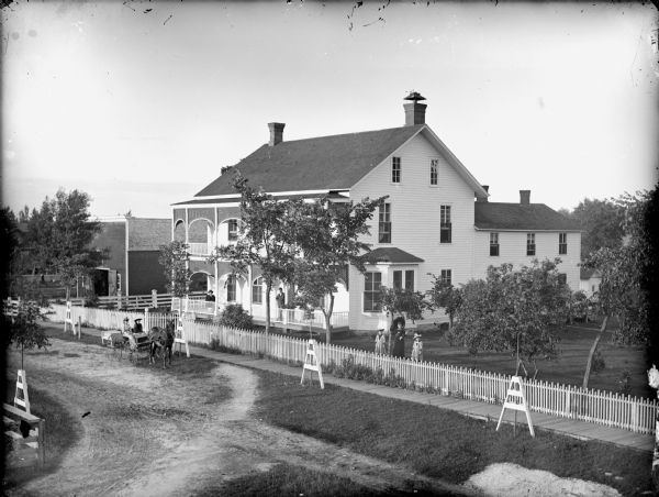 Elevated view of a woman and two girls posing standing in a yard behind a fence. On the road on the left are two women sitting in a buggy pulled by a single horse on a road, and two men are standing and sitting on the porch of the large two-story frame house, probably the H.A. Bright house.	