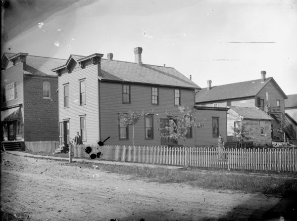 View across road of a man and woman posed standing in the fenced yard of a house in town, which is to the right of a storefront of the I.O.O.F. (International Order of Odd Fellows) 294. A dog is sitting on the stoop of the house.	