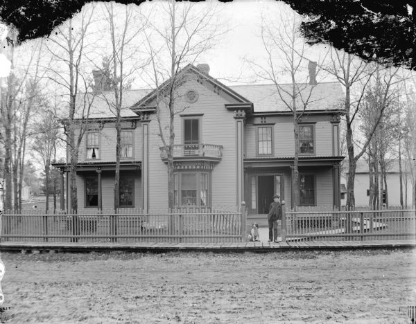 View across road of a boy and dog posed standing at the open gate of a fence in front of a large house. Probably the green house owned by Henry Ormsby on the northeast corner of Sixth and Tyler Streets, which later burned down.	