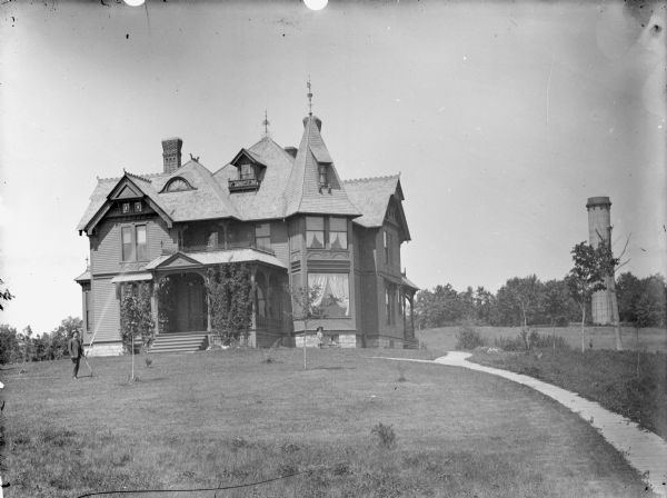 View across lawn of girl posing standing with small baby carriage, and a man posing standing and watering the lawn. They are in front of a Victorian-style frame house, probably the residence of W.R. O'Hearn located at Tenth and Harrison Streets, which was later set on fire by A.J. McNab. There is a tower in the background on the right.