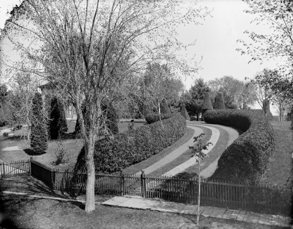 Elevated view of curved footpath behind a fence. The path is lined with hedges and trees and leads to a large house. Probably the grounds surrounding the Spaulding residence.	