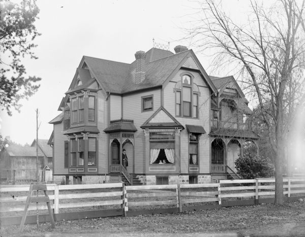 Exterior view of a frame house. A man is posed standing on the small side porch on the left, and two children are posed sitting looking out of one of the front windows. Possibly the residence of L.C. Jones, torn down in 1994.