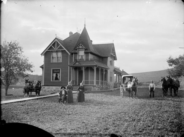 View of group of people posing on the lawn in front of a two-story frame house, possibly the residence of Hans Kjorstad. On the left is a man sitting in a buggy pulled by a team of two horses, in the center a man is sitting in a chair near two women who are standing. On the right two children are sitting in a buggy pulled by a single horse, and on the far right a man is standing near a team of two horses. A dog is lying on the porch in the background.