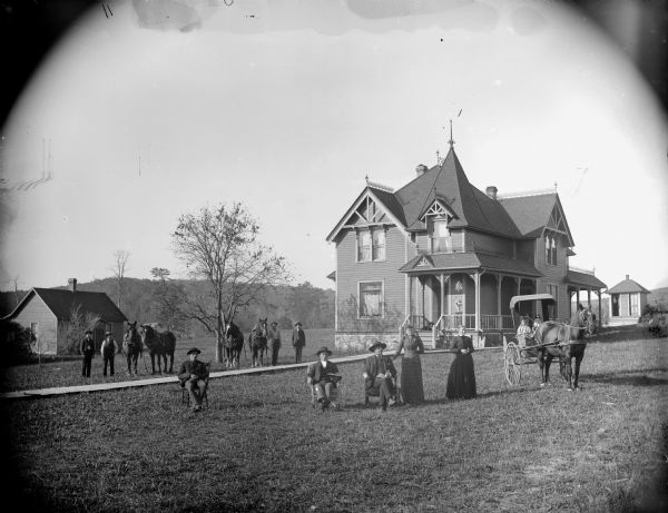 View of group posing on the lawn in front of a two-story frame house. Four men in the background on the left are standing near two teams of horses; three men are sitting in chairs on the lawn near and two women are standing; on the right are two children sitting in a horse-drawn buggy. Possibly the residence of Han Kjorstad. A dog is lying on the porch in the background.