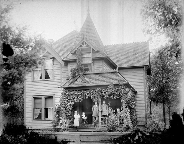 Group of people posing on the porch of a two-story frame house. Possibly the residence of Han Kjorstad. On the porch is a young girl standing and holding a doll, a boy sitting on a tricycle, two men standing, and a woman sitting.