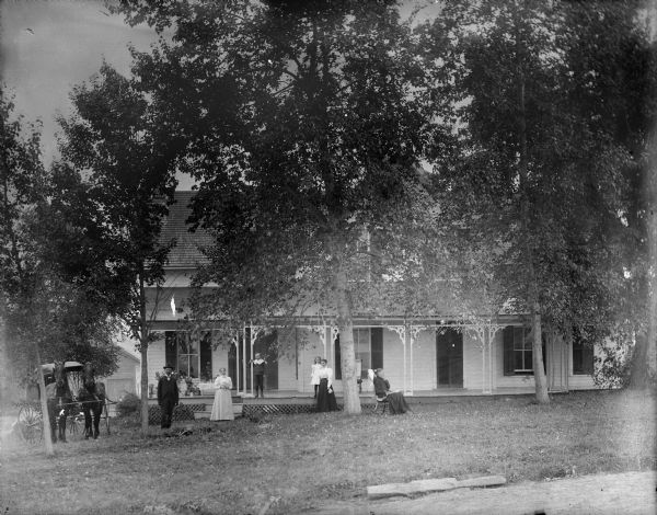 View across lawn towards a group of people posing in front of a two-story frame house. There is a woman sitting in a rocking chair, two women and a man are standing, a boy and girl are on the porch, and on the left is team of horses with a buggy. In the background beyond trees is a barn.