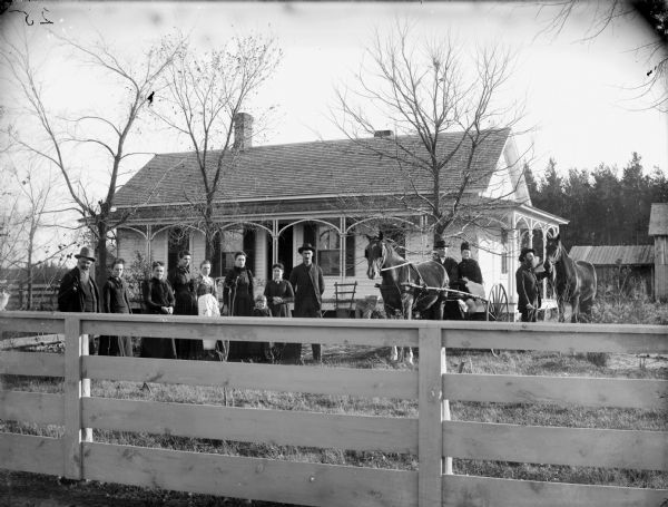 Two men, six women, and a young girl are posing standing in the yard in front of a single-story frame house. There is also a man and woman sitting in a buggy pulled by a single horse, and a man beside them displaying a horse.