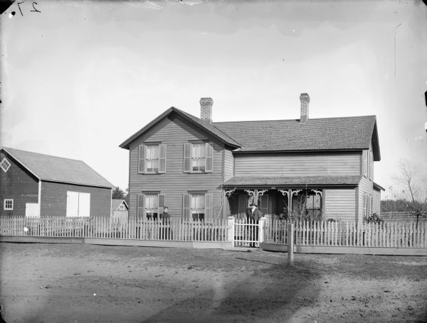 View across road towards a man and woman posing standing behind a picket fence in front of a frame house. The man appears to be holding a cane in his left hand, and is using a crutch under his right arm.